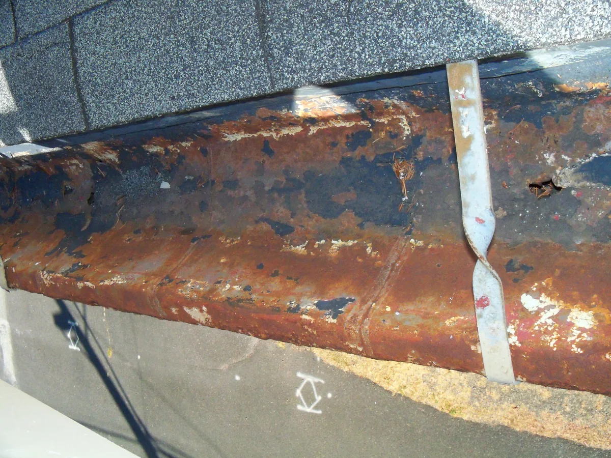 Rusted metal gutter with visible holes beneath overlapping roof shingles.